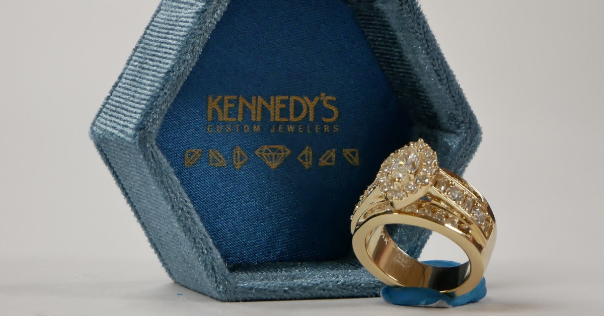 A gold ring with clear stones standing beside an open blue velvet jewelry box on a light gray surface.